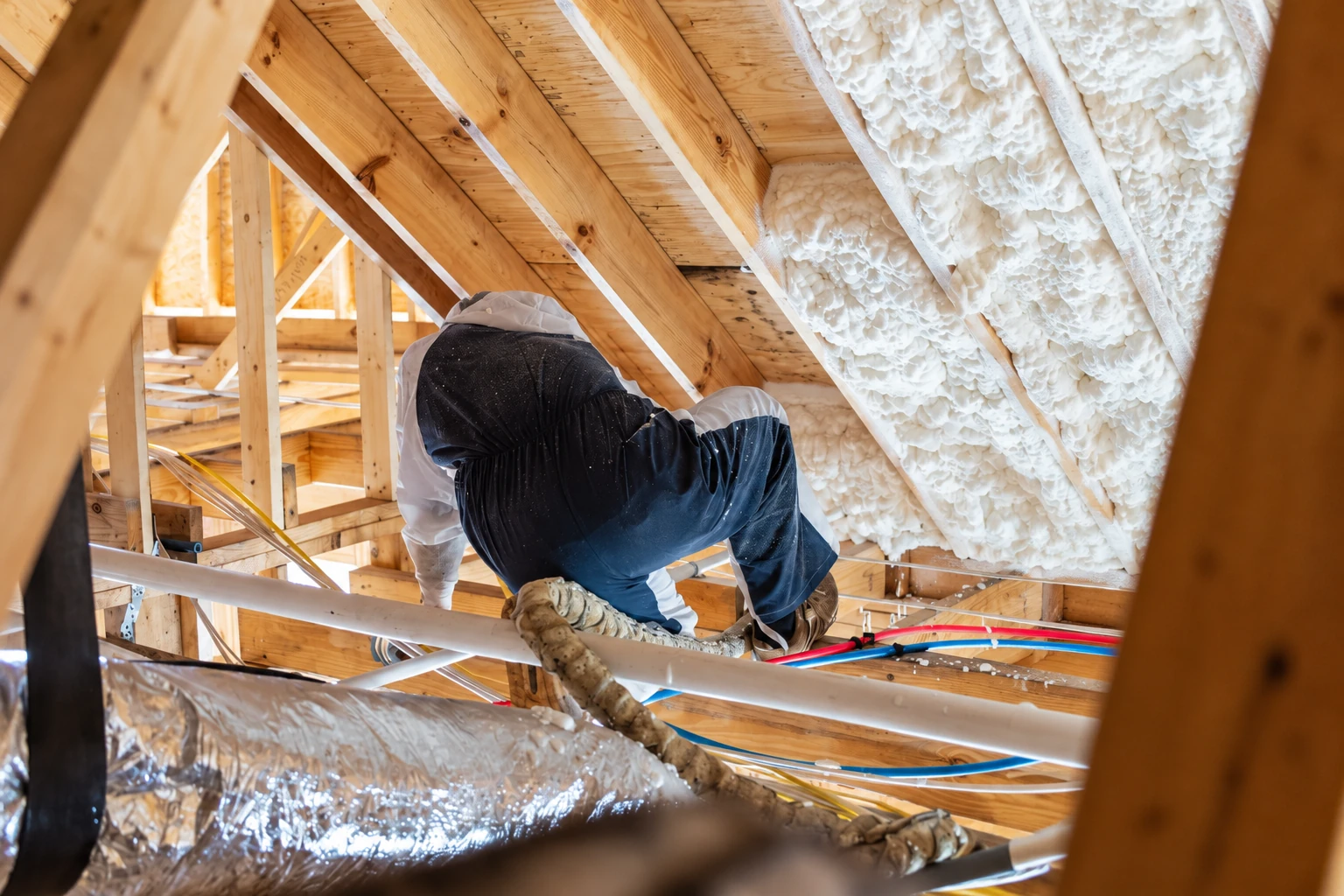Insulation worker kneels among attic framing beside densely applied foam insulation on the sloped roof.