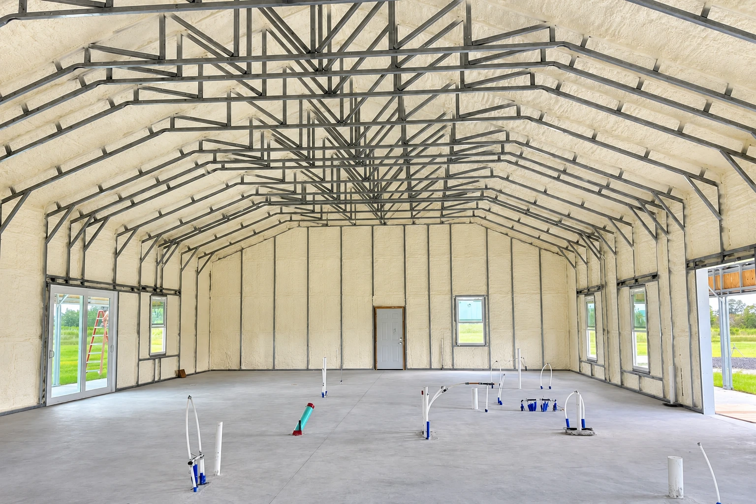 A large commercial building interior has pale spray foam coating its walls and roof beneath exposed steel trusses.