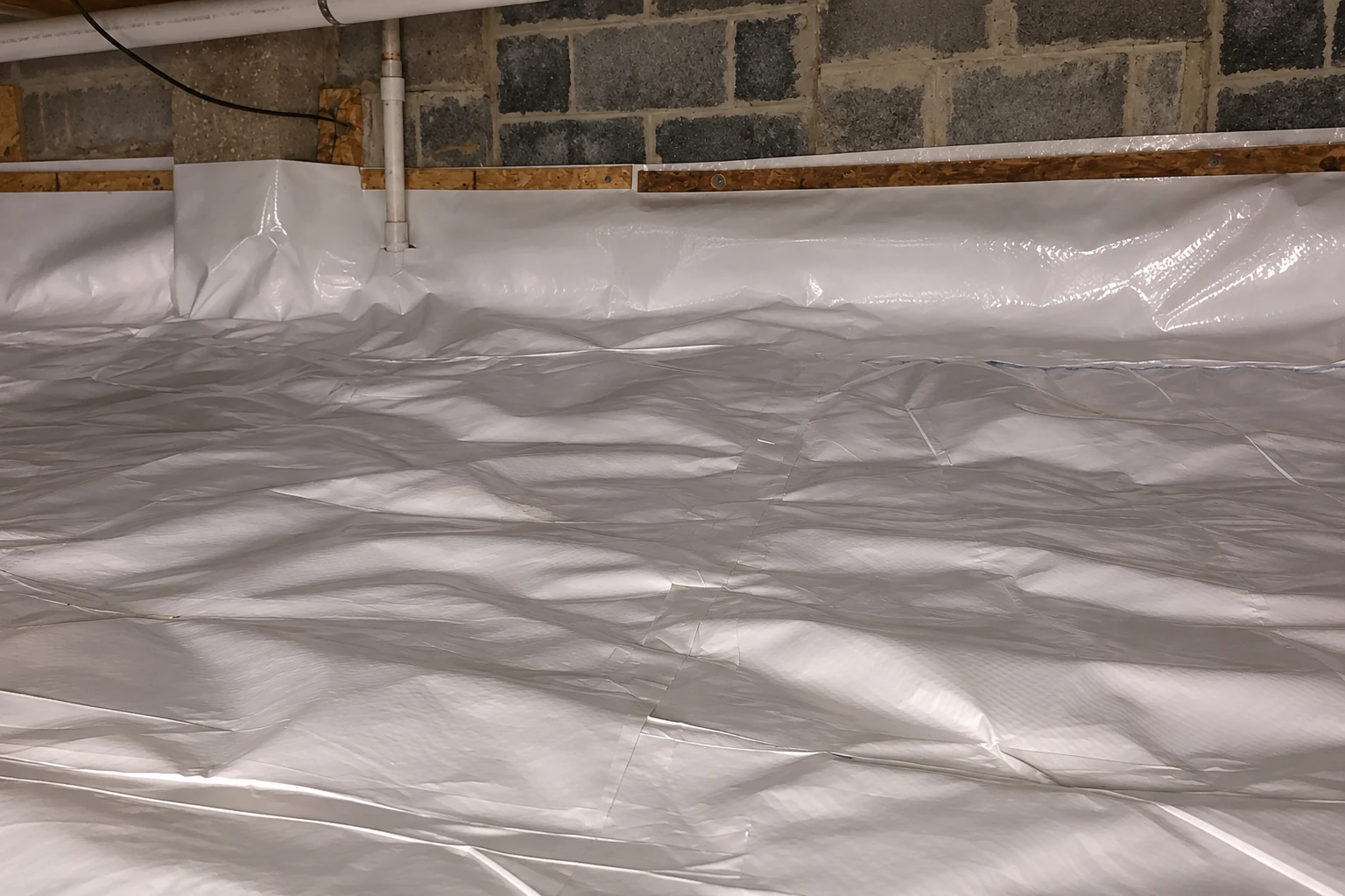 A crawl space has white plastic vapor barrier spread across the floor and wrapped along the lower block walls.