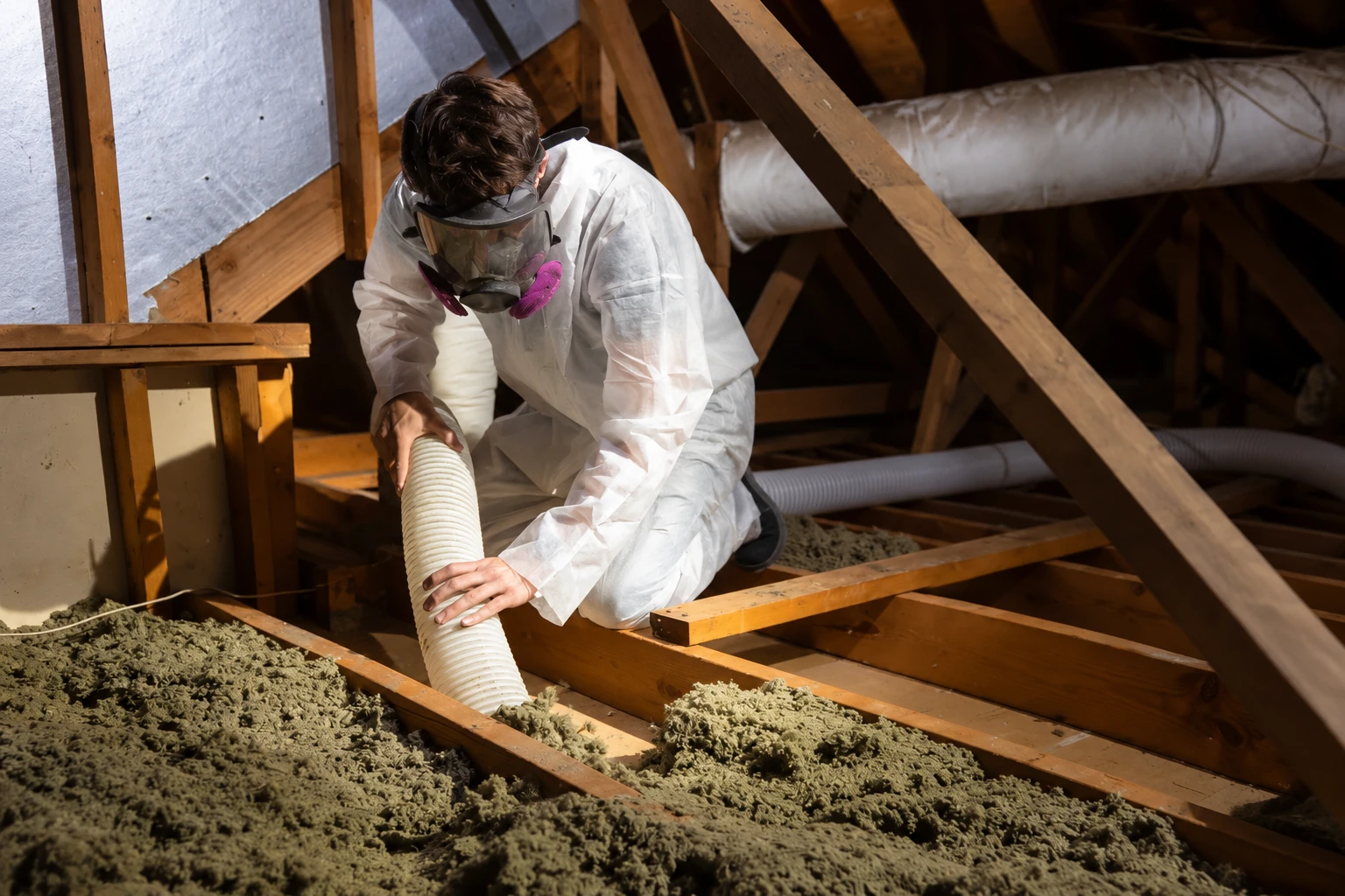 A worker in protective coveralls and a respirator vacuums loose attic insulation through a hose.