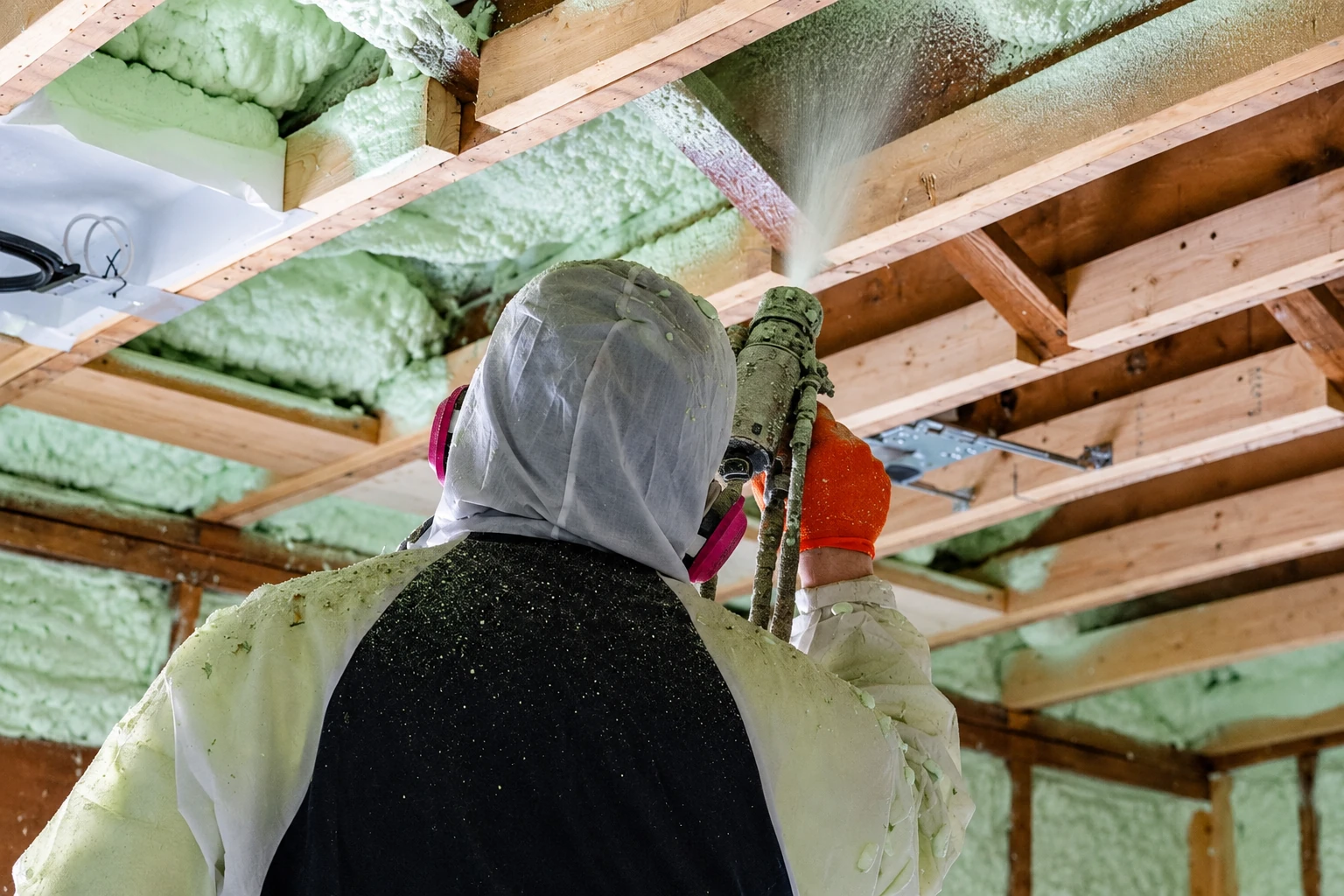 A worker in protective gear sprays pale green foam insulation between exposed attic roof rafters.