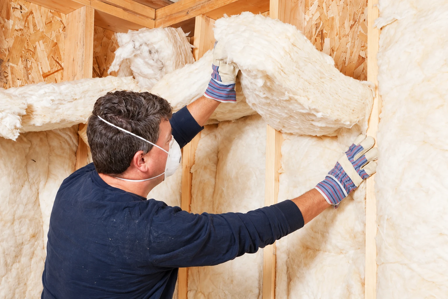 A masked worker fits fluffy white insulation between wooden wall studs.